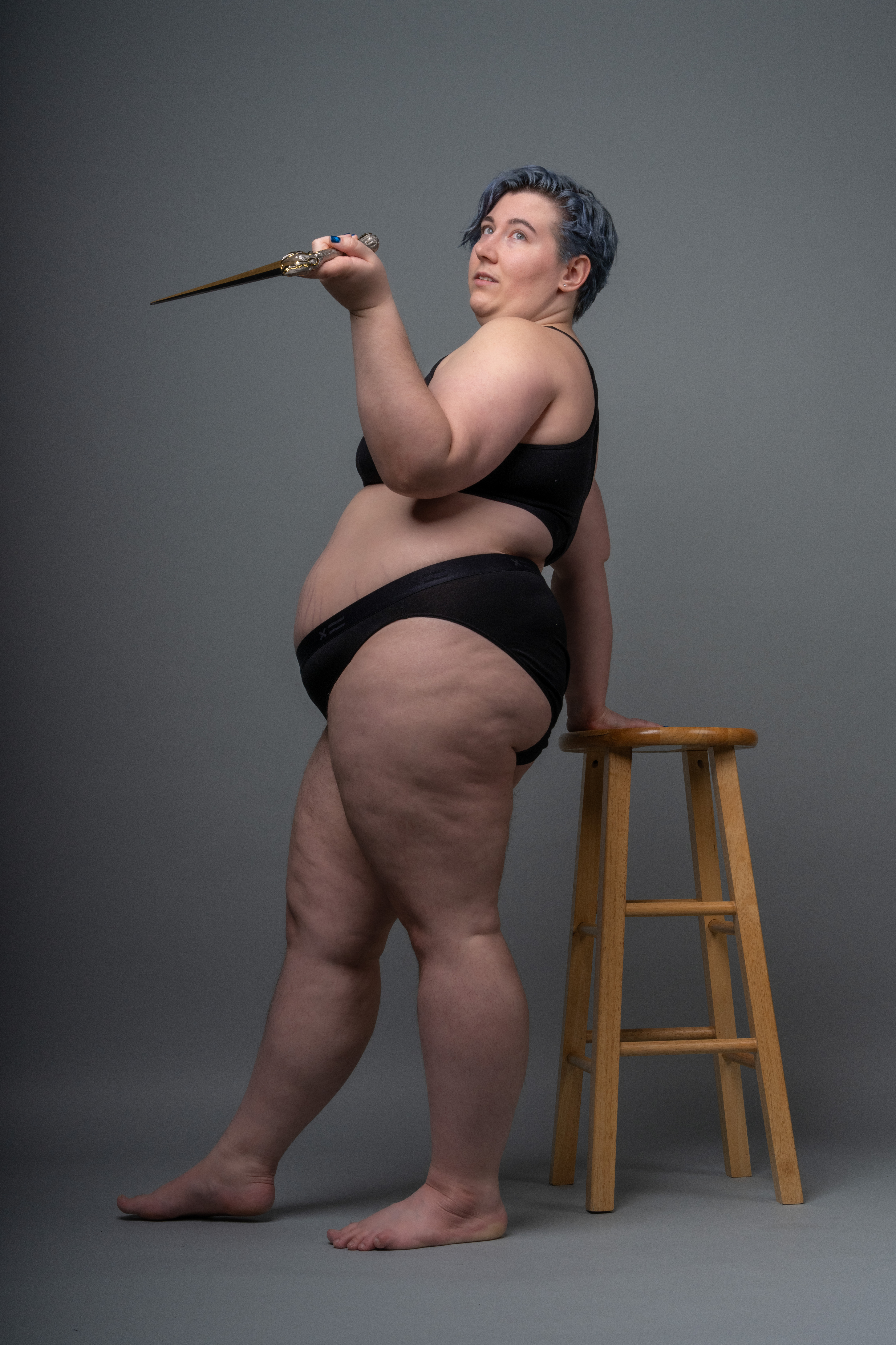 a plus size woman in black underwear stands in front of a grey background with one hand resting on a wooden stool and holding up a wand in the other hand. she is looking up towards the wand with a curious expression. she has blue hair.