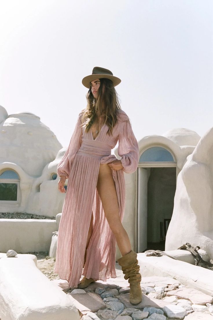 a woman in a long pink dress and cowboy hat walks along a stone walkway in front of a white building. the setting appears to be desert like.