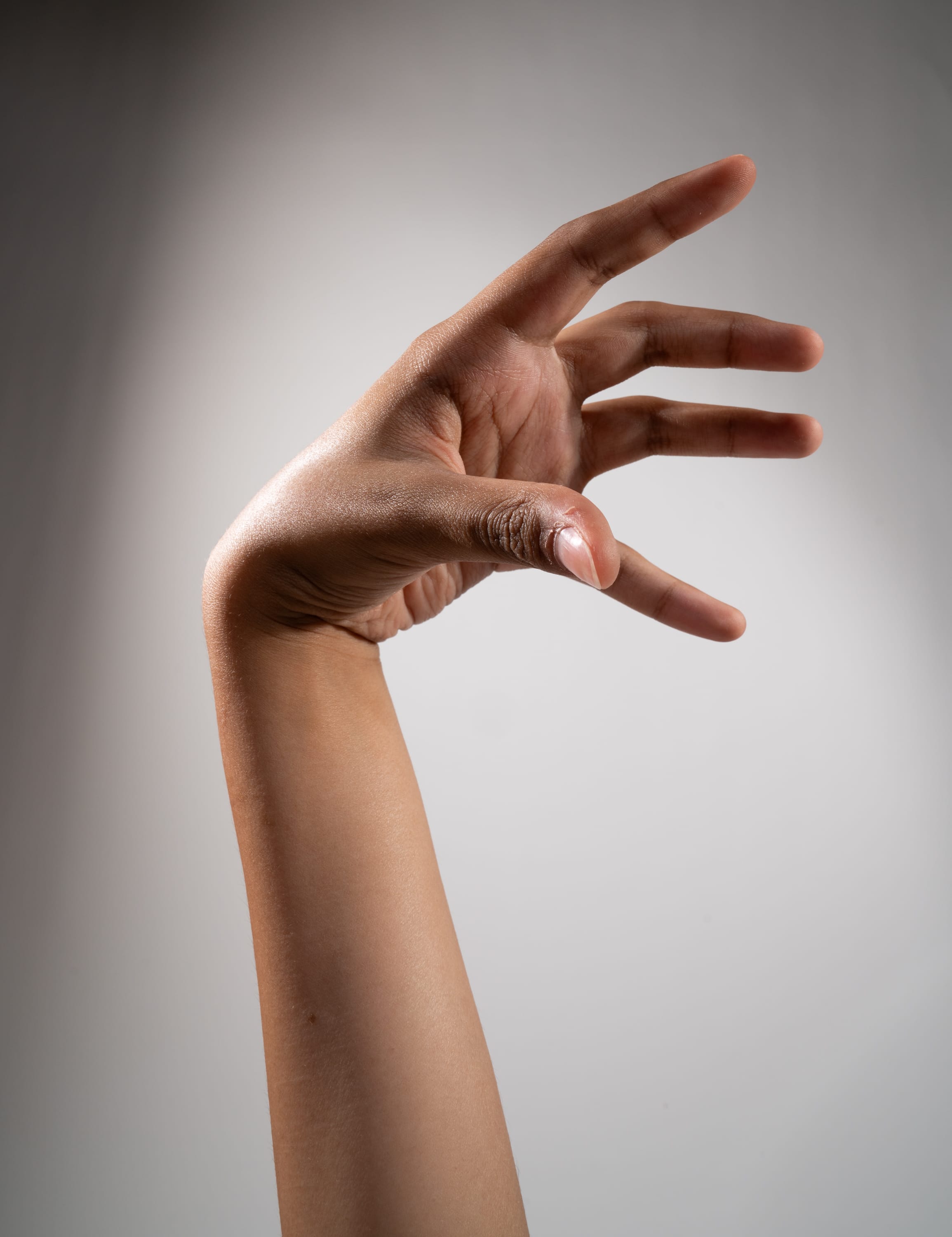 a single hand and arm of a person with brown skin is reaching out against a plain gray background. the hand is open with the fingers slightly spread as if reaching to grab something. the image is lit with studio lighting creating a soft gradient on the background.