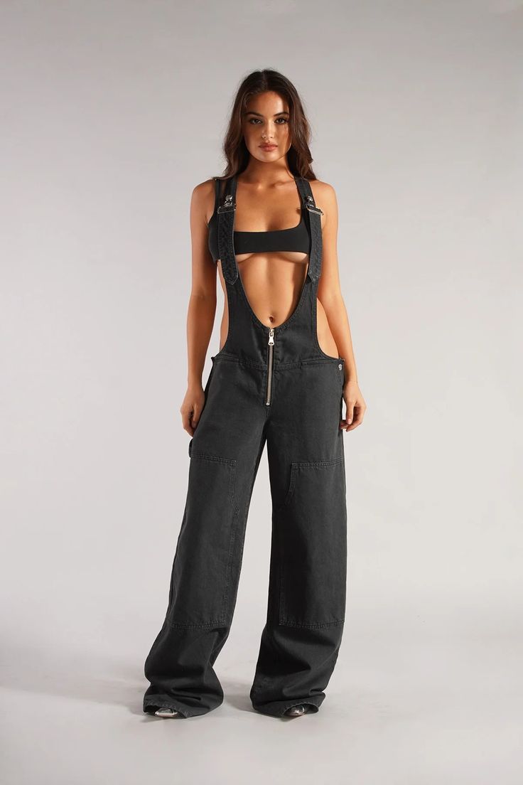 a woman is standing in a studio, wearing black wideleg overalls with a front zipper and cutouts at the sides. the overalls are styled with a black bandeau top. the background is a light gray, and the lighting is soft.