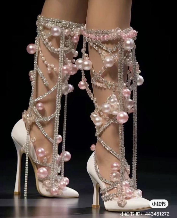 loseup shot of a pair of exquisite high heels adorned with an abundance of pearls and crystals. the shoes are white with intricate detailing, featuring delicate chains and dangling pearl accents. the heels are tall and slender, and the overall aesthetic is one of luxury and sophistication.  the shoes are shown on a model's legs on what appears to be a fashion runway. a small watermark is visible in the bottom right corner.