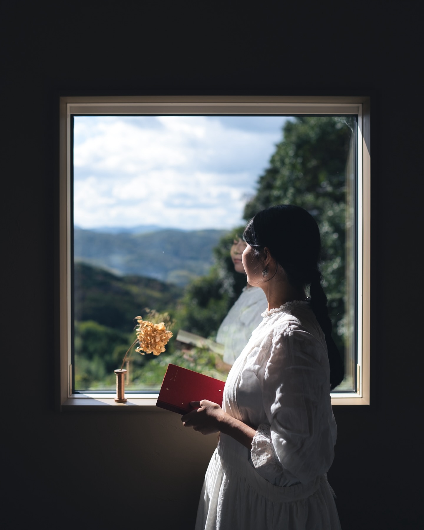 a young woman stands by a large window overlooking a picturesque landscape of rolling green hills and mountains. she holds a red book in her hands and gazes out the window, appearing peaceful and contemplative. the scene is bathed in soft, natural light.  she is wearing a white dress. a small, dried flower arrangement sits on the windowsill. the overall mood is serene and tranquil.