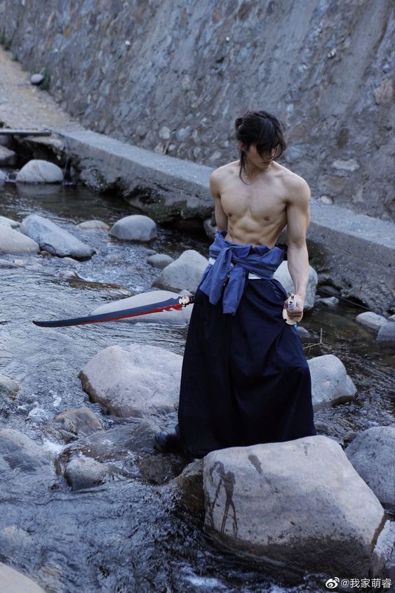 a muscular man, shirtless with a blue cloth tied around his waist and a black skirt, stands in a flowing stream with a large katana sword beside him. he is cosplaying a samurai warrior and has long black hair tied back. the environment is natural and rocky, with the image showcasing a serene and powerful aesthetic.