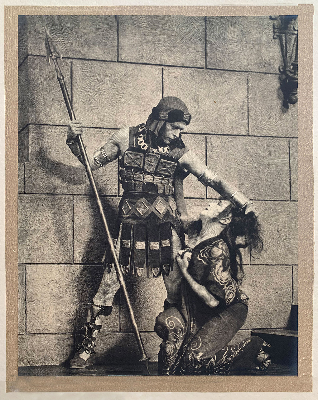 the image is a striking black and white photograph, possibly a vintage print or a reproduction of one, depicting a dramatic scene from a theatrical production.  it appears to be set in a classical or ancient setting, suggested by the brick wall backdrop and the costumes. a male actor, dressed as a warrior or soldier in what looks like ancient greek or roman attire, complete with a breastplate,  holds a spear and stands over a kneeling female actor. the woman is dressed in flowing robes; her expression and posture suggest distress or fear. the overall mood is intense and dramatic, highlighting the characters’ interaction. the high contrast and detailed textures within the image are characteristic of vintage photography techniques. the background is a simple brick wall, focusing attention on the main subjects in the foreground.