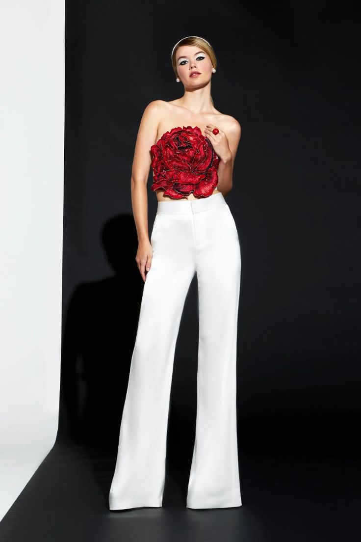 a fashion model is posing in a studio shot. she is wearing a white pantsuit with a red flower top. her hair is pulled back and she is wearing minimal makeup. the background is black and white.