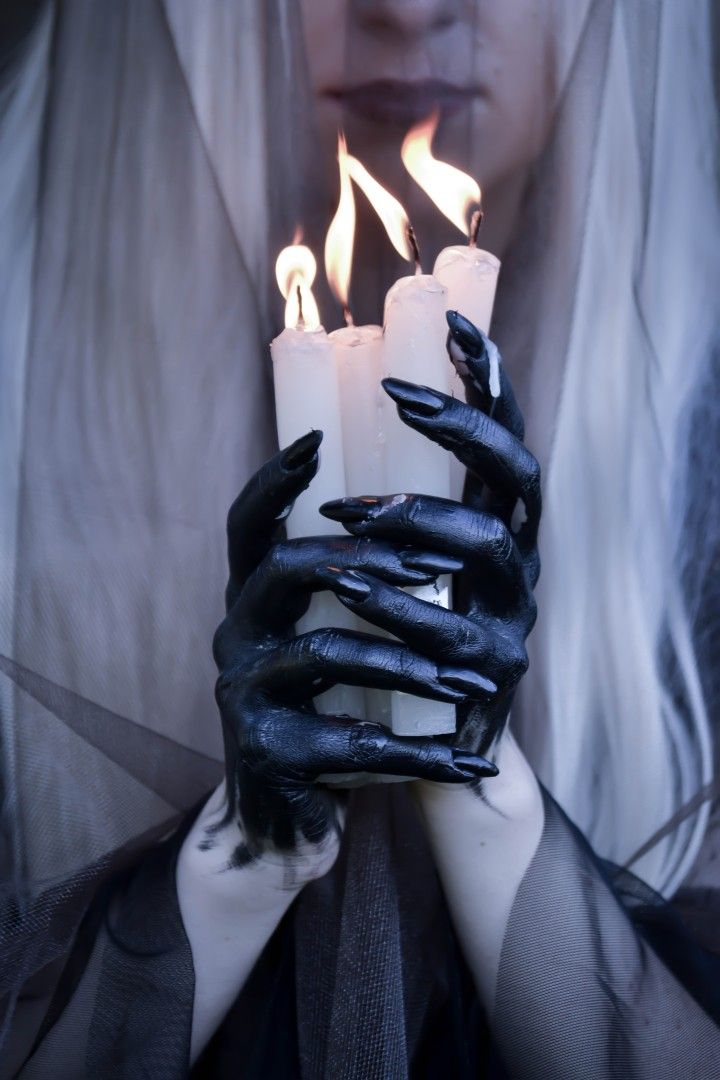 the image showcases a closeup view of a woman's hands, painted black with long, sharp nails, delicately holding a cluster of burning white candles.  the woman's face is partially obscured by a sheer, lightcolored veil. the overall aesthetic is gothic and mysterious, with a focus on dark, dramatic lighting and a sense of occult or supernatural themes. the contrast between the dark hands and the bright flames of the candles is striking. the image evokes feelings of mystery and intrigue.