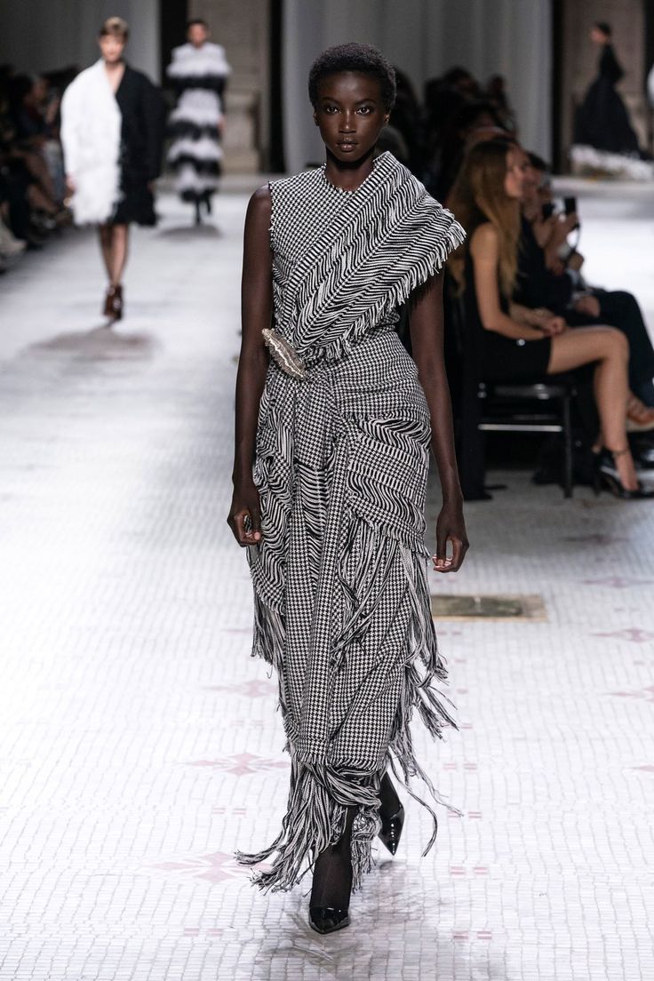 a black woman model walks down a runway wearing a long, elegant black and white gingham dress with fringe detailing. the dress has a draped design and a belted waist. the model is wearing black high heels. the background shows other models and spectators. the overall setting suggests a high fashion runway show.