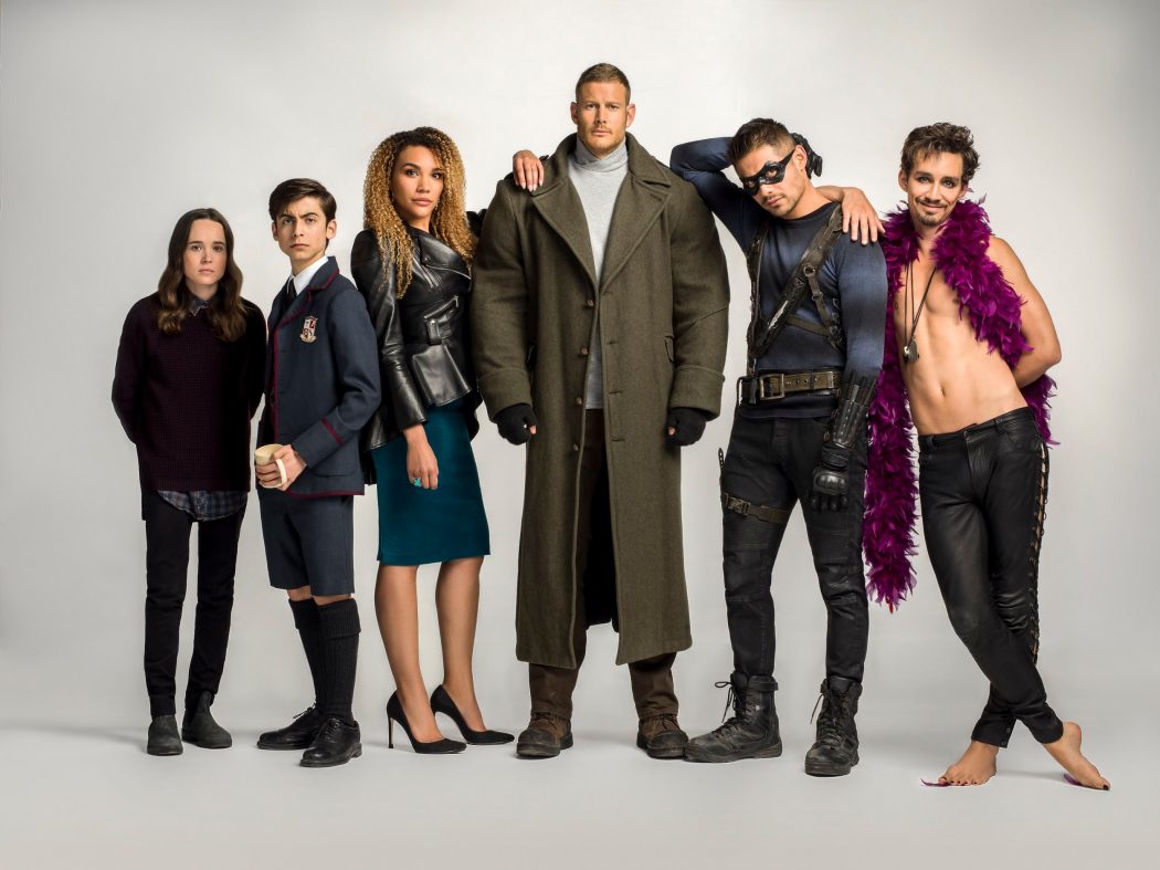 a promotional image for the netflix series "the umbrella academy" featuring the six main cast members standing in a line against a white background. they are all wearing costumes reflecting their character's personalities. from left to right: number seven (page), number five,  allison, luther, diego, and klaus.