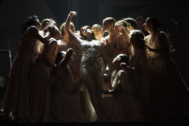 a dramatic stage performance featuring florence welch of florence + the machine, centrally positioned and surrounded by a chorus of female dancers.  florence is dressed in a shimmering, sequined gown, and the dancers are in flowing, lightcolored robes. the lighting is dramatic, highlighting florence and creating a mystical and powerful atmosphere. the overall image evokes a sense of otherworldly beauty and theatrical grandeur.