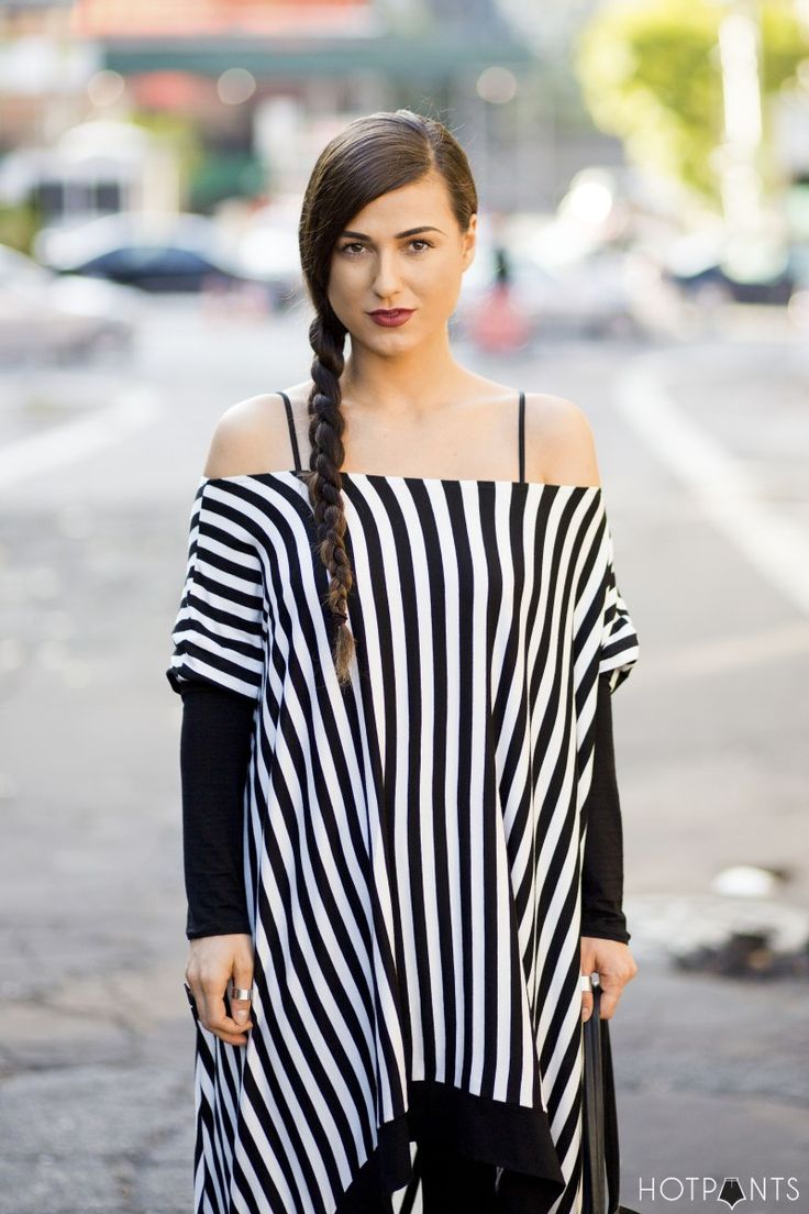 a woman stands facing the camera wearing a black and white striped offtheshoulder top over a black longsleeved shirt. her hair is styled in a braid down one side, and she wears dark lipstick. she is standing on a street.