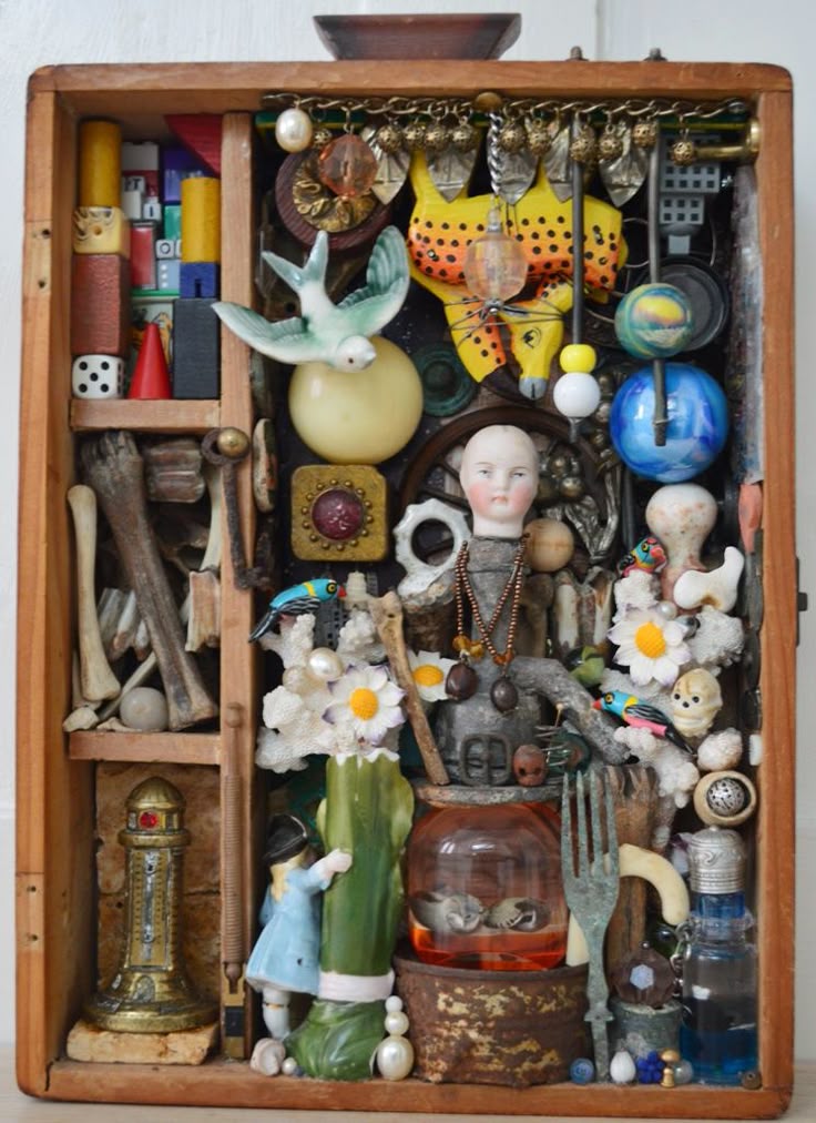 this is an assemblage art piece housed within a wooden box that resembles a curiosity cabinet. it features a collection of diverse found objects and vintage items. the contents include a doll head, a bird figurine, bones, a die, a doll figurine, keys, vintage toys, trinkets, found materials, beads, spoons, forks, bottles, miniatures, skulls, a lighthouse replica, coral, flowers, and a yellow butterfly. the mixed media artwork presents a combination of textures and shapes, demonstrating creative expression through folk art and outsider art styles. the overall effect is a unique and handcrafted art object.