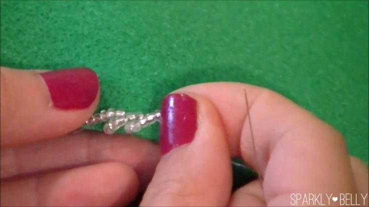 a closeup shot shows hands with red nail polish doing beadwork on green fabric. the hands are holding a strand of clear beads and using a needle to sew them onto the fabric. the image is cropped tightly around the hands and the craft being performed.