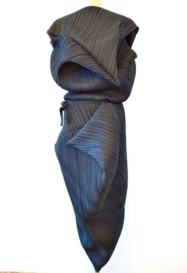 a closeup of a pleated dress in a dark shade of grey, made with many folds and pleats. the dress is draped over a mannequin and is displayed in a white room, showing off the intricate detail of the garment. the light and shadow play on the pleated fabric creates an interesting and dynamic effect.