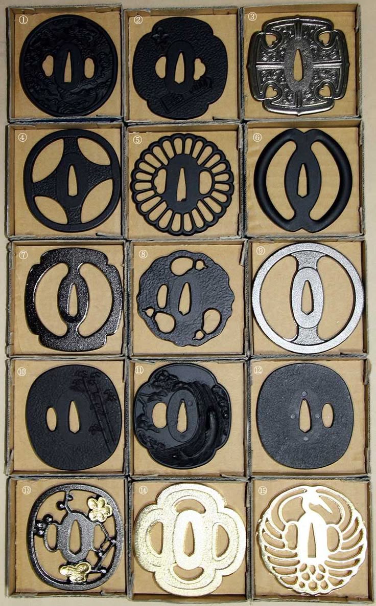 the image displays a collection of fifteen sword guards (tsuba), typically used on japanese swords such as katanas. they are arranged in a 3x5 grid within individual cardboard boxes, suggesting they might be part of a collection or for sale. the tsuba vary in design, color, and material, ranging from black and silver to gold, with intricate patterns and motifs. some are circular, while others have more elaborate shapes. these decorative pieces serve to protect the user's hand and add aesthetic value to the sword.