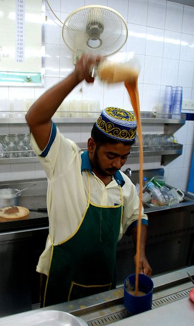 a closeup shot of a man in a cafe, wearing a green apron and a kopiah (traditional muslim hat), expertly pouring teh tarik (pulled tea).  the tea is being poured from a highheld glass into a lower cup creating a long, thin stream. he's standing in a stainless steel kitchen area with stacks of glasses visible in the background. the focus is on the action of pouring the tea, highlighting the skill and technique involved in making this popular malaysian drink. a ceiling fan is also visible in the background.