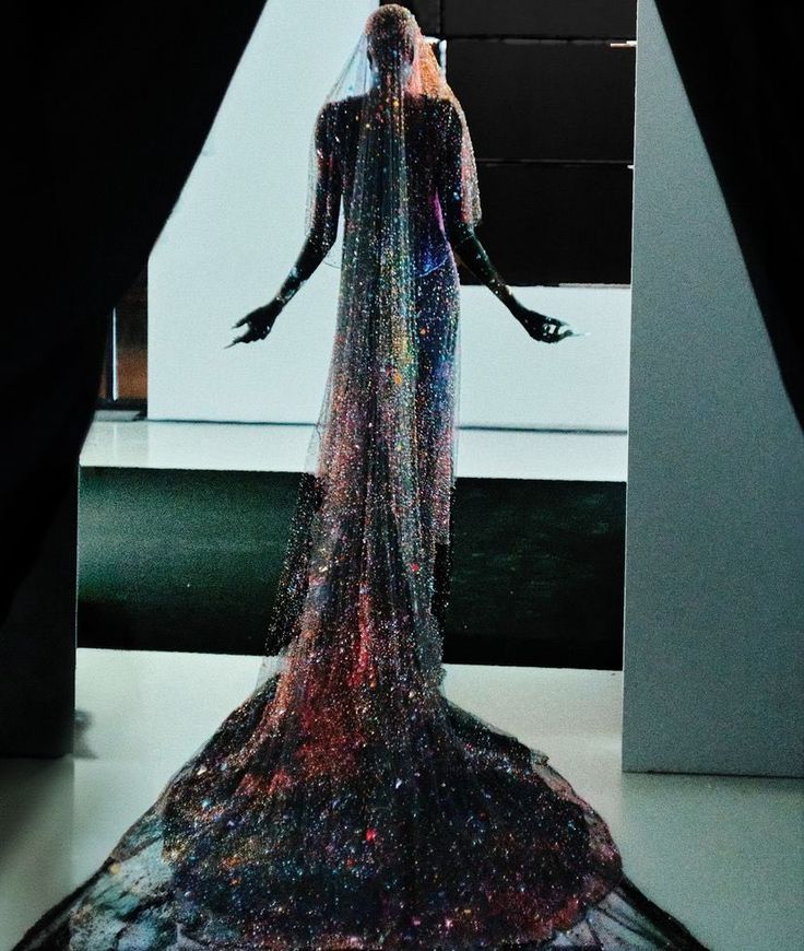 a mannequin is on display wearing a long, sparkling gown with a trailing veil. the gown appears to be covered in glitter or sequins, creating a celestial or ethereal effect. the color scheme features a mix of dark and light shades, with the glitter adding pops of color. the mannequin is positioned in front of a stark black and white backdrop, which further enhances the dramatic and formal presentation.