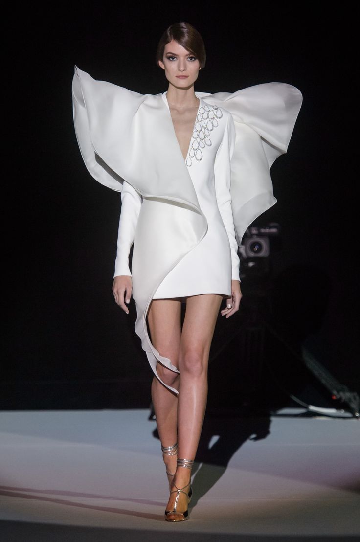 a fashion model walks the runway wearing a  white, longsleeved dress with a dramatic, ruffled shoulder detail. the dress is  embellished with  jewelry.  the model is wearing  strappy heels  and has her hair pulled back in a sleek style. she is striking a pose for the cameras.