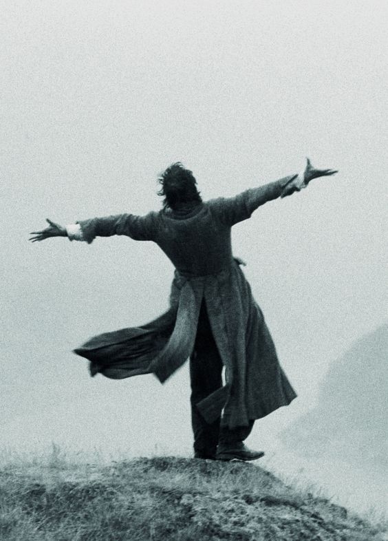 a black and white image of a person standing on a hilltop with their arms outstretched. they are wearing a long coat that is billowing in the wind. the sky is overcast, creating a sense of drama and solitude. the overall palette consists of varying shades of gray, emphasizing the somber atmosphere.