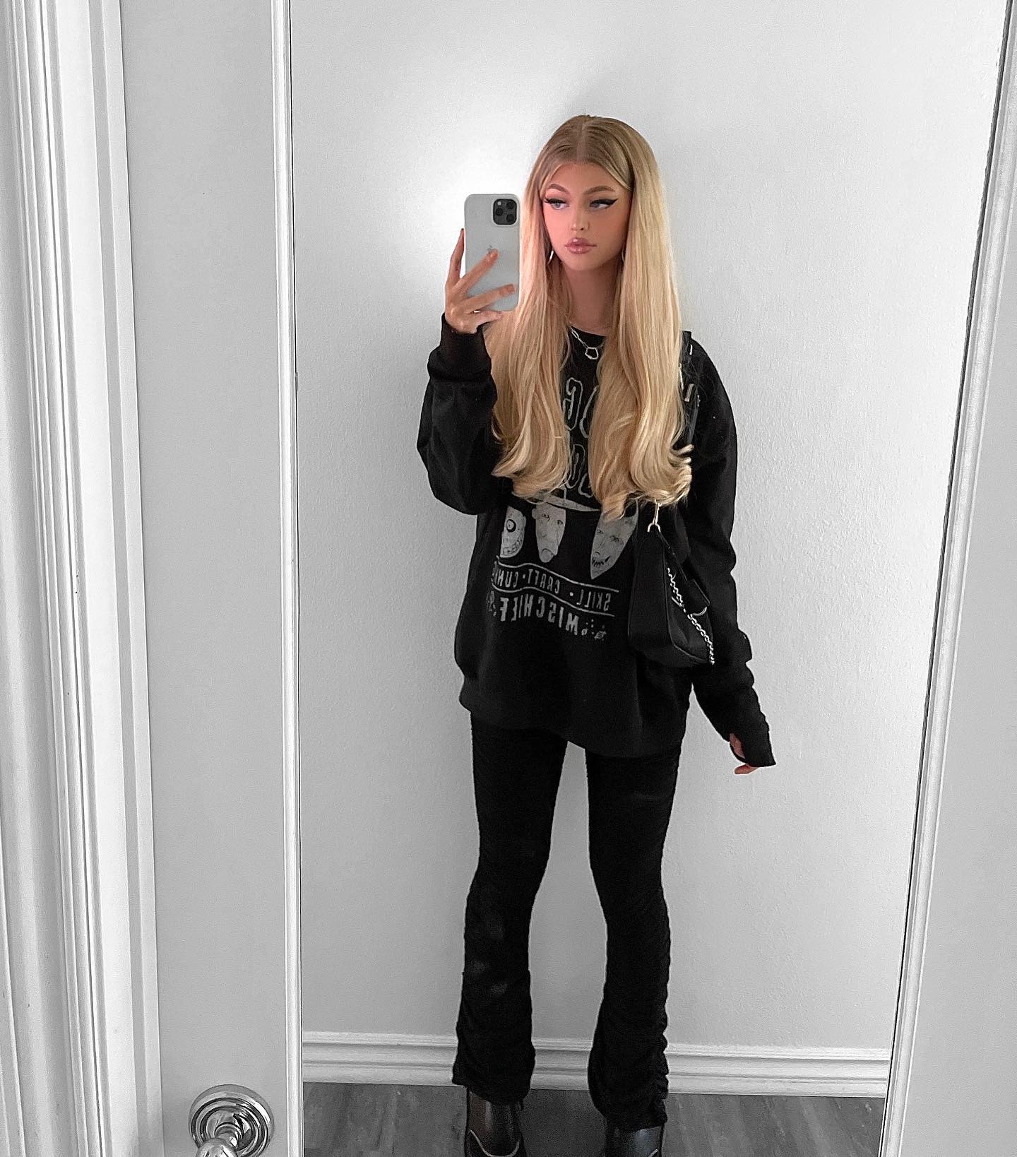 a woman with long blonde hair is taking a mirror selfie. she is wearing a black sweatshirt with a graphic design, black leggings, and black boots. she has a black purse slung over her shoulder and is holding a cell phone in her hand. the woman is standing in front of a white wall with a door on the left and a mirror on the right.