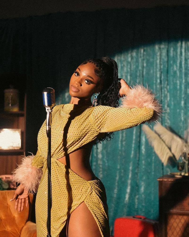 a woman wearing a sequined yellow dress with fur accents is singing into a microphone on a stage. she has her hand on her hip and is looking down at the microphone. the background is a teal colored wall.