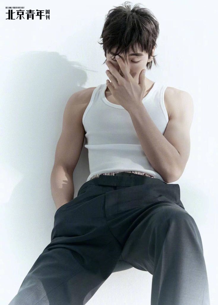 a man is sitting on a white background, wearing black pants and a white tank top. he is looking down and has one hand on his face. the other hand is in his pocket. the man is wearing a lot of rings on his fingers.