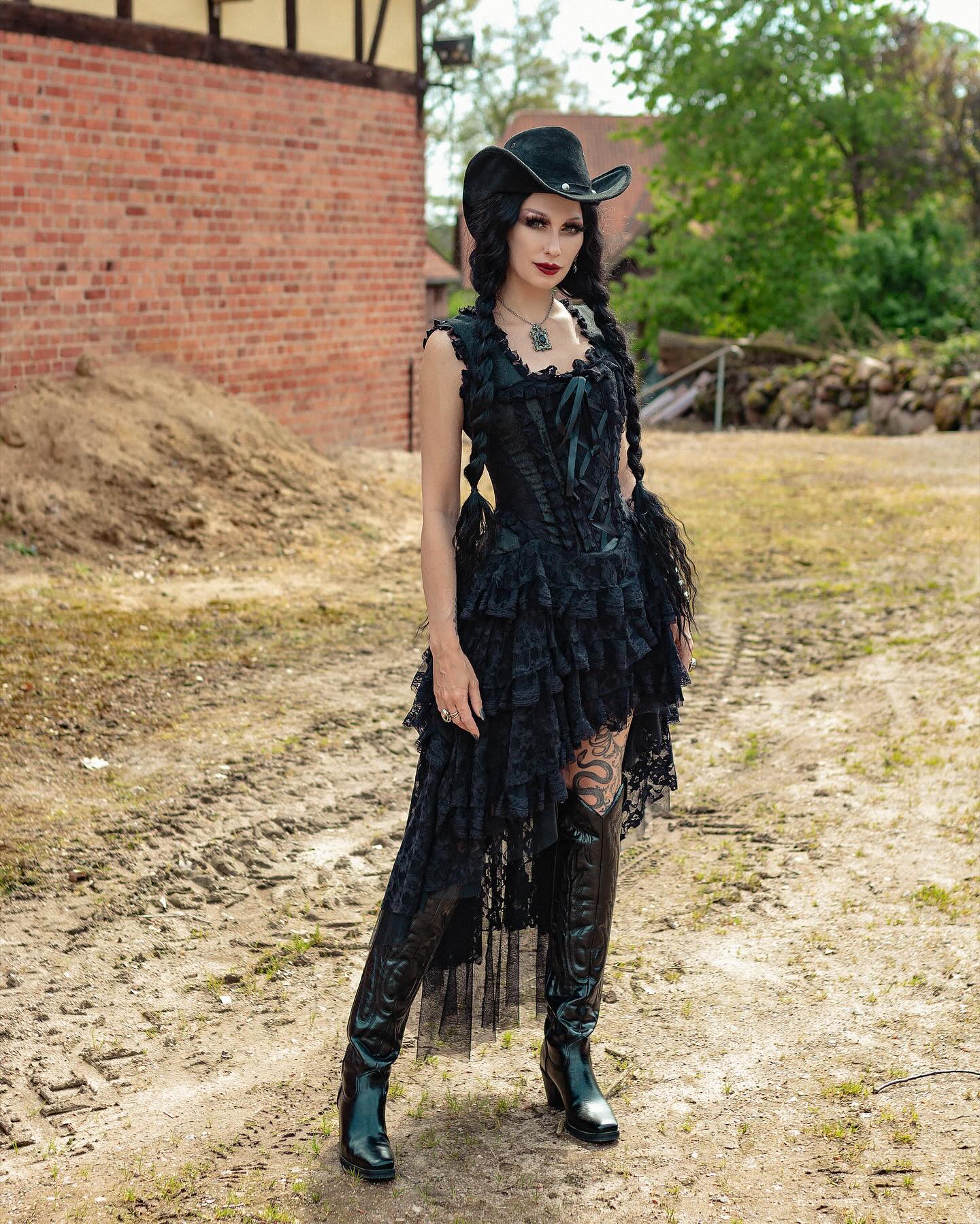 a full shot of a gothic girl in a black lace dress and cowboy hat. she is posing outdoors on a dirt road with a brick wall in the background. she is wearing high black boots.  her hair is in long braids. she has a corset bodice and a highlow hemline.  the style is a mix of gothic lolita and western.