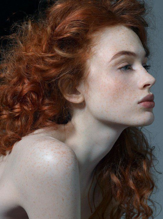 the image is a closeup, artistic portrait of a young woman with vibrant, curly red hair and numerous freckles across her pale skin.  she is shown in profile, her head slightly tilted, her gaze directed away from the camera. her shoulders and neck are exposed, highlighting the texture of her skin. the lighting is soft and subtle, emphasizing the ethereal quality of the subject and her features. the overall aesthetic is elegant and natural.