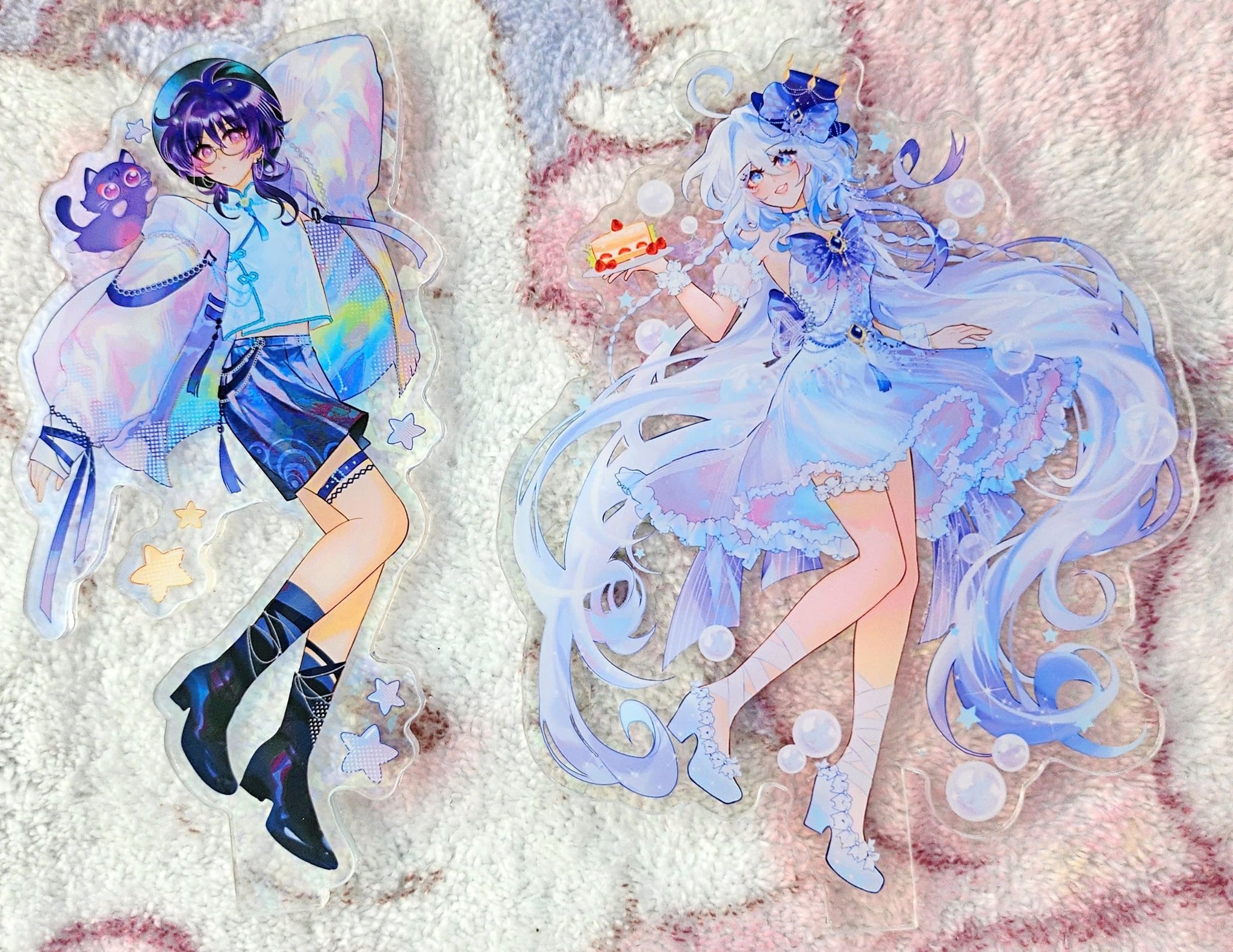 he image shows two acrylic keychains featuring animestyle characters.  one keychain depicts a boy with dark purple hair wearing a light blue and white outfit with dark blue shorts and boots. he's accompanied by a small dark purple catlike creature. the other keychain showcases a girl with long white hair styled with a light blue bow. she's dressed in a light blue pastel dress and is holding a small cake. both keychains have a transparent acrylic background with added decorative elements like stars and bubbles, enhancing their pastel and magical atmosphere.  the keychains are placed on a soft pink fabric background.