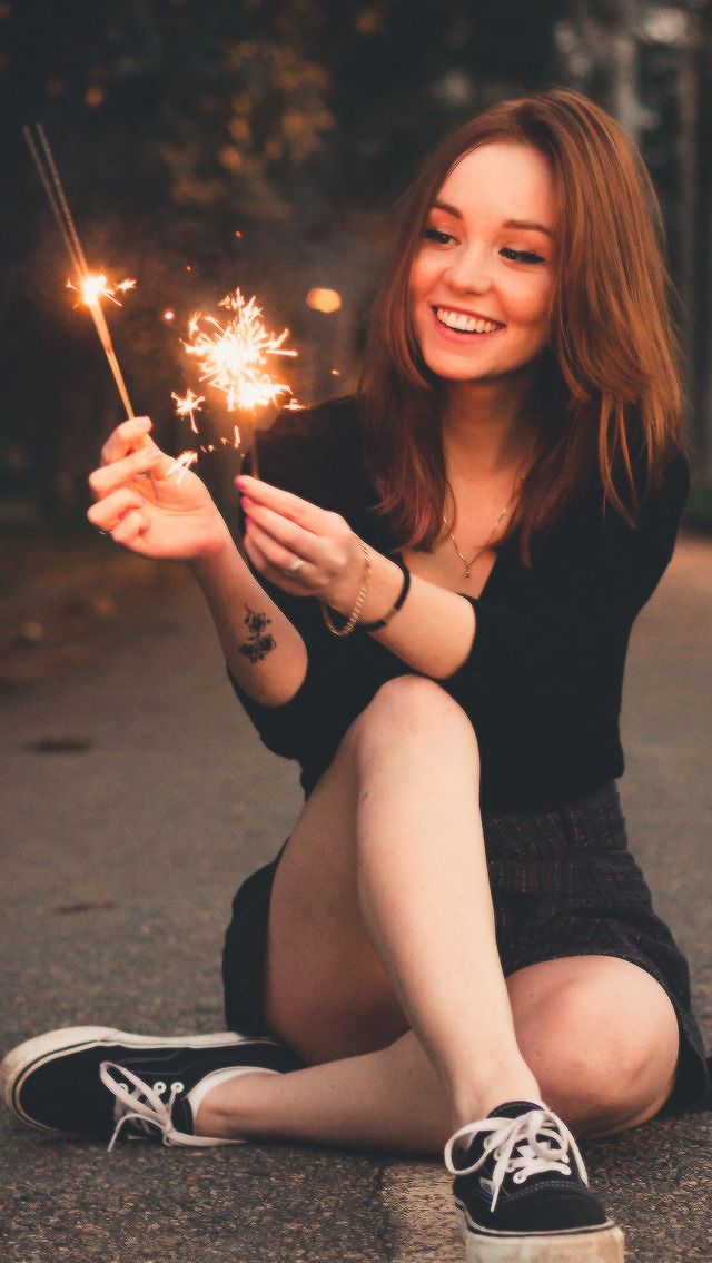 a young woman is sitting on the street at night, smiling brightly as she holds two lit sparklers. she is wearing a black longsleeved shirt and a black and white checkered skirt, paired with black and white sneakers. the warm glow of the sparklers illuminates her face and creates a joyful and celebratory atmosphere. the background is dark and blurred, drawing focus to the woman and the sparklers.