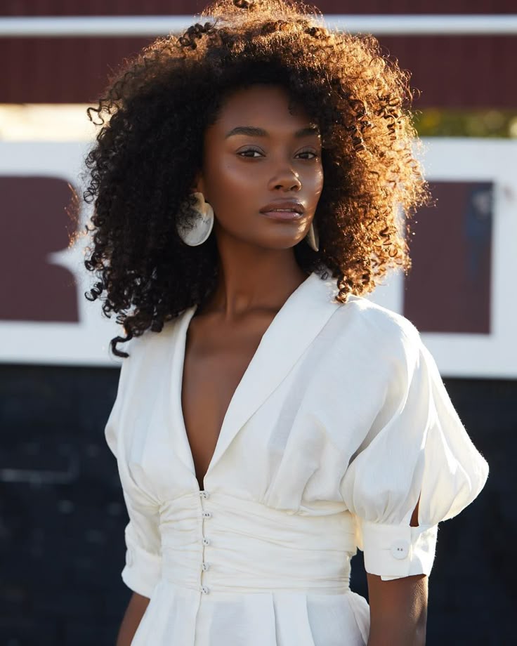 a portrait of a woman with dark skin and curly hair, wearing a white vneck dress with puff sleeves. she is wearing large, round earrings. sunlight illuminates her hair and face. the background is a dark and light colored wall.