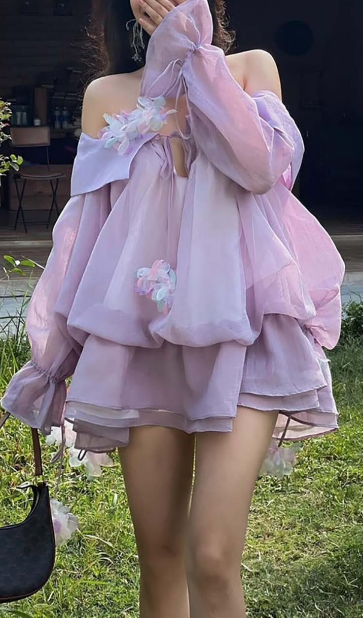 a young woman wearing a beautiful light purple dress with flowers. she is standing in a grassy area with a gentle smile on her face.  her dress has ruffles and a bow at the neck, and she is accessorizing with a black handbag. she has a very soft, girly and feminine style.