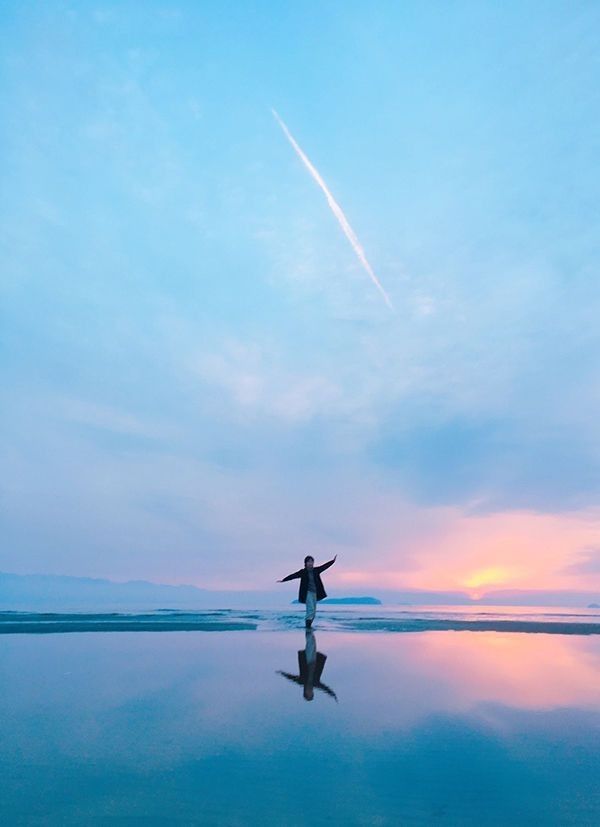 a lone woman stands on a calm, reflective beach at sunset, her arms outstretched. the sky is a blend of pastel pink, orange, and blue hues, creating a serene and picturesque scene. the woman's reflection is perfectly mirrored in the still water, enhancing the sense of tranquility and balance.  a contrail streaks across the sky adding another layer of interest to the scene. the overall atmosphere is peaceful, dreamy, and evokes feelings of freedom and serenity.