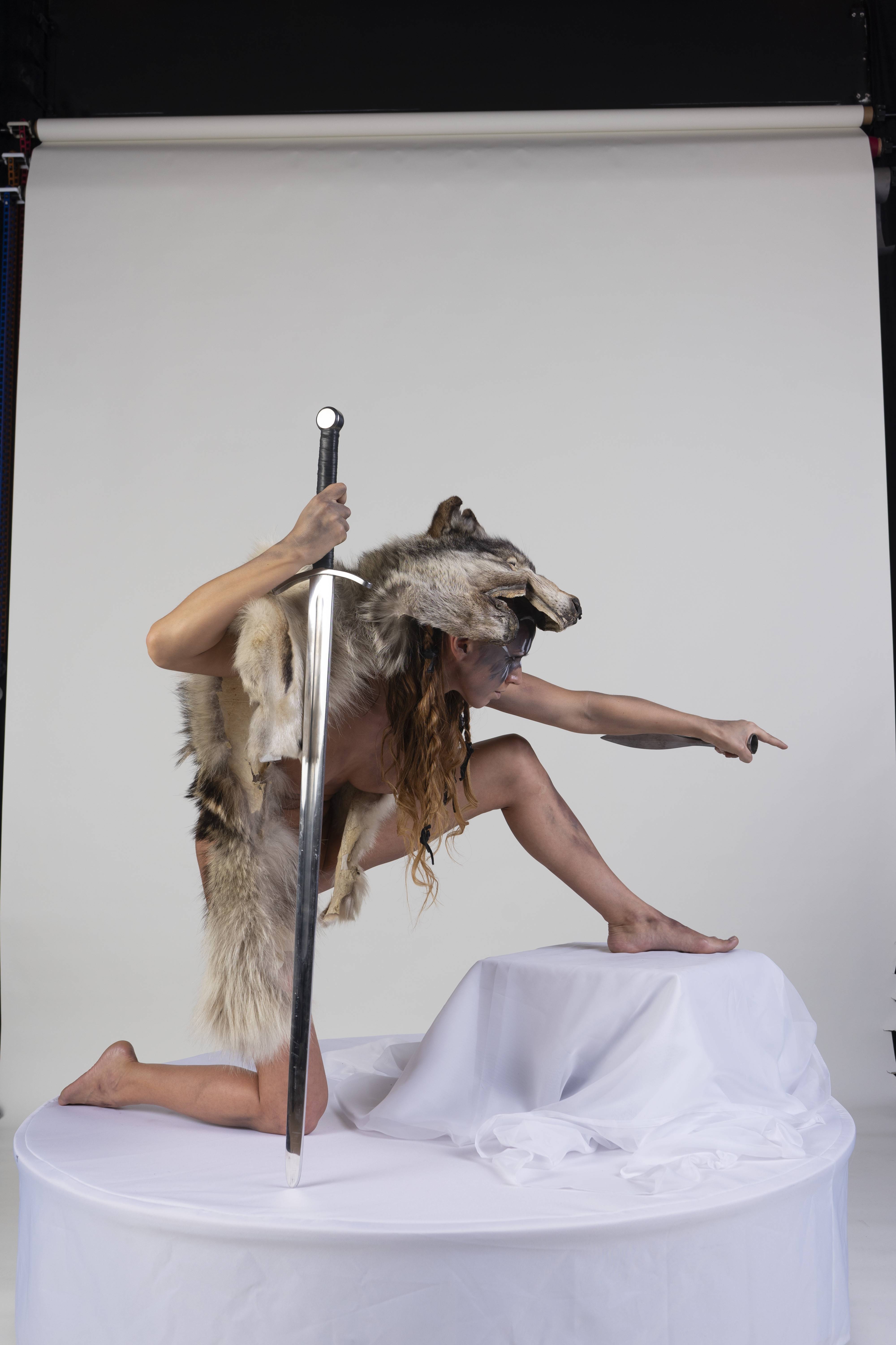 a woman wearing a wolf skin and holding a sword is kneeling on a white platform. she is pointing to the right with her right hand. the background is a plain white studio backdrop. the woman appears to be in costume, possibly for a fantasy or historical themed shoot.
