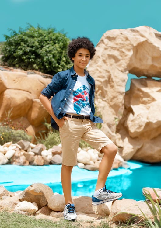 a teenage boy with curly brown hair is standing by a small pool in a rocky outdoor setting. he's wearing a white tshirt with a graphic design, a dark blue buttondown shirt, light beige shorts, and gray and blue sneakers. he's smiling and looking at the camera with his hands in his pockets. the background features large rocks and lush greenery. the overall atmosphere is relaxed and casual. the image seems to be a fashion photograph showcasing casual summer attire for teens.