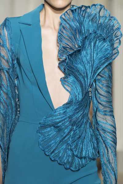 a woman wearing a luxurious, teal blue gown with intricate, sparkly ruffles and embroidery. the dress features a plunging neckline, a fitted bodice, and flowing sleeves. the ruffles are layered and cascading, creating a dramatic and elegant look. the fabric has a subtle shimmer and sheen, making it look even more luxurious. the woman's hairstyle and makeup complement the dress, creating a chic and sophisticated overall style.