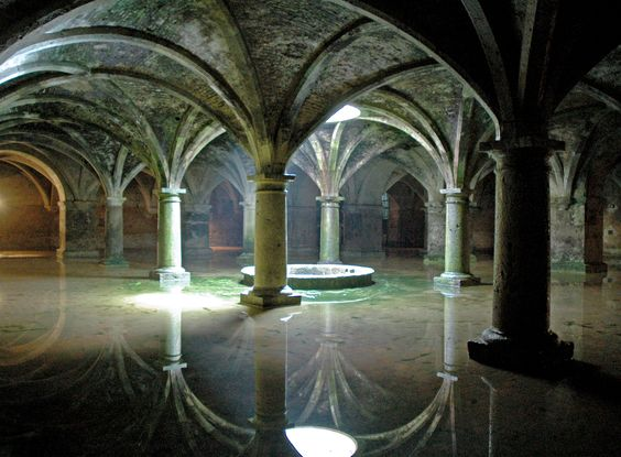 the image depicts an ancient underground cistern filled with water. tall stone pillars with arched vaults support the ceiling, their reflections visible in the water. the stone structures are covered with moss, and light shines through openings in the ceiling, creating reflections and illuminating parts of the dark space. a well is visible in the middle of the cistern.