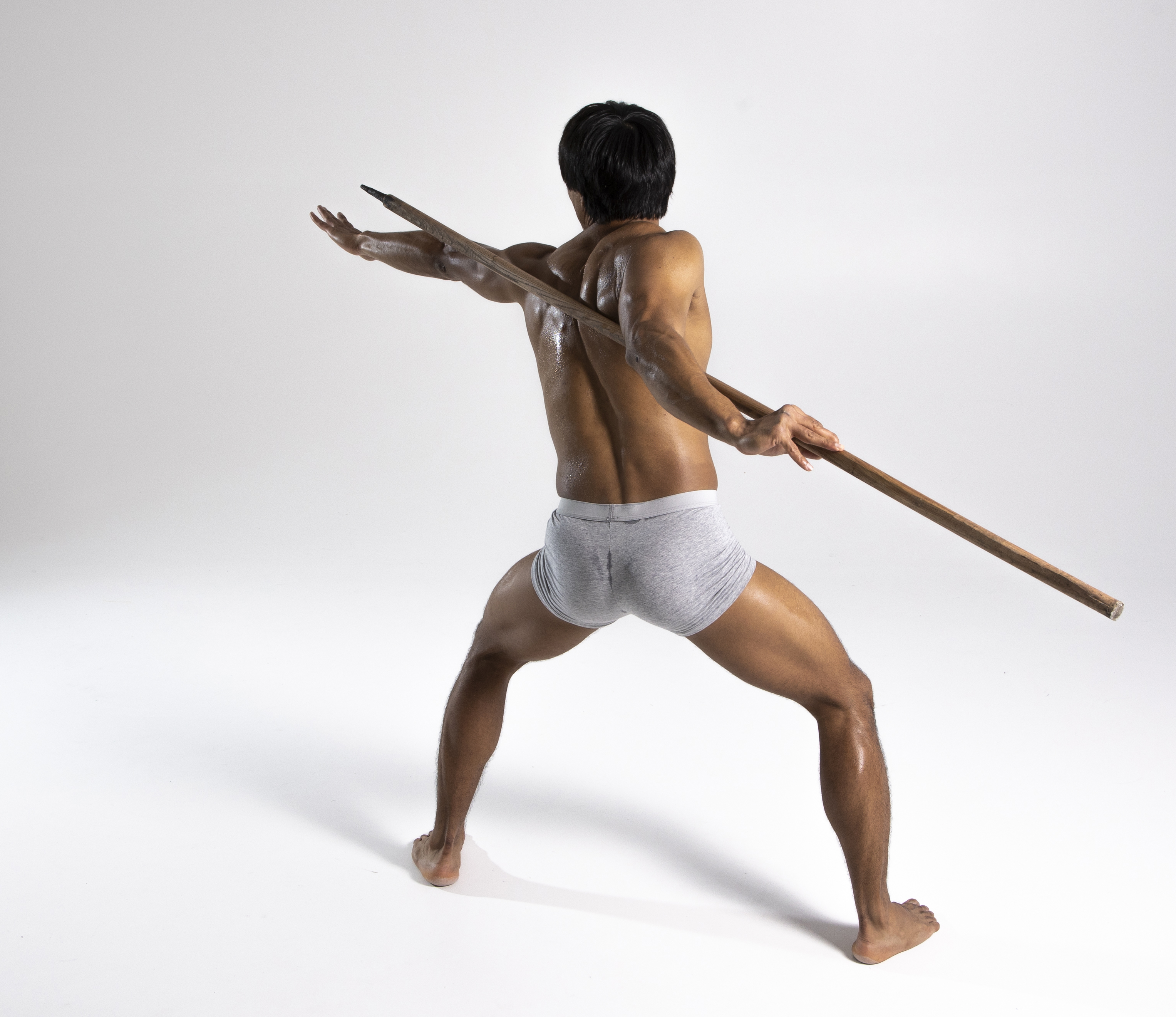 a shirtless man, seen from the back, is holding a spear in a defensive stance. he's wearing underwear and is barefoot in a studio setting.