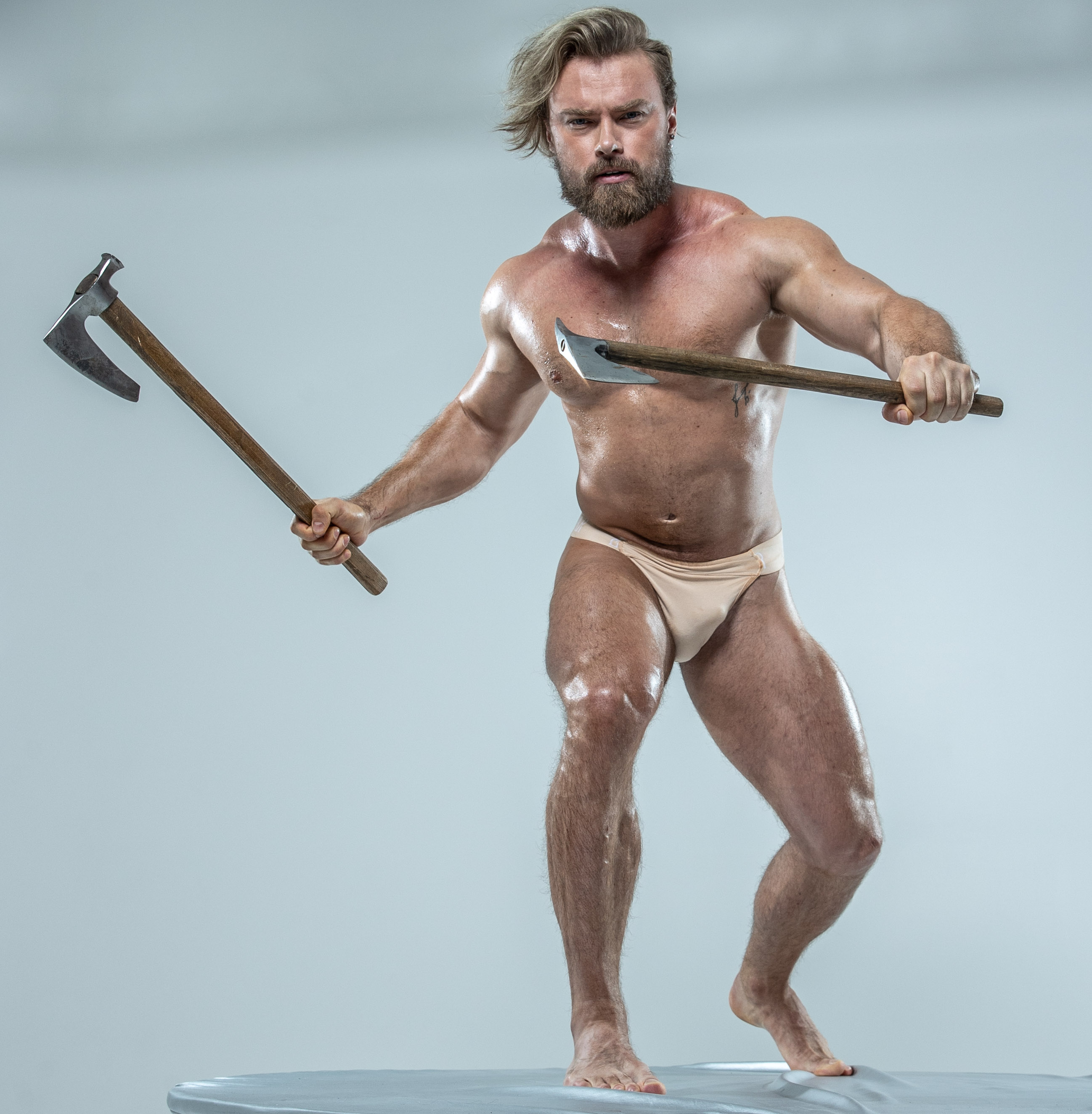 a muscular, bearded man stands in a fighting stance against a grey background. he holds aloft two axes, one in each hand, ready to strike. he is barechested, wearing only white underwear, showcasing his physique. his expression is intense, conveying strength and aggression. the lighting is bright, highlighting his muscles and the sharp edges of the axes. the overall impression is one of power, masculinity, and a connection to history or mythology.