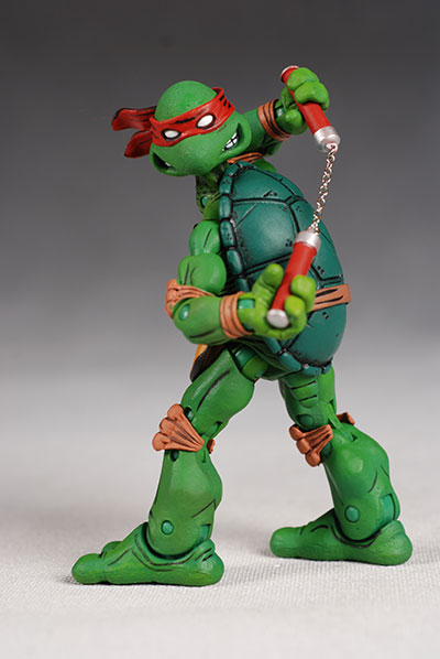 a teenage mutant ninja turtle raphael action figure stands on a light surface. he wears a red bandana and wields nunchucks. his shell and body are shades of green, with brown wrist and ankle bands.