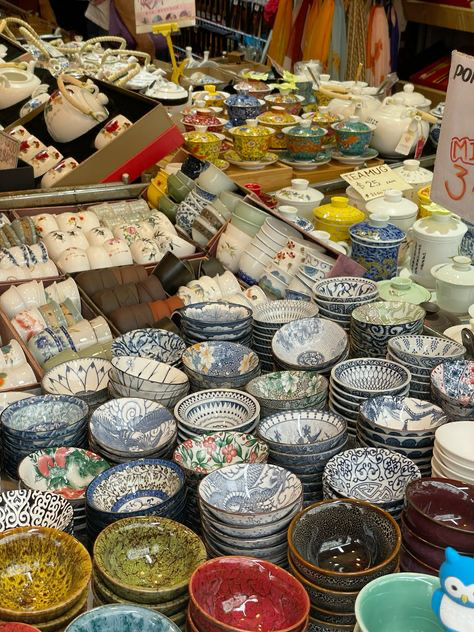 the image shows a vibrant display of various teacups, tea bowls, and other teaware items in a market stall or shop. the items are predominantly porcelain or ceramic, showcasing a range of colors, patterns, and styles.  many are stacked neatly in piles, while others are individually displayed.  the setting appears to be an asian market, given the style of the teaware and the overall atmosphere.  the image suggests a large selection of traditional and possibly handmade items available for purchase.