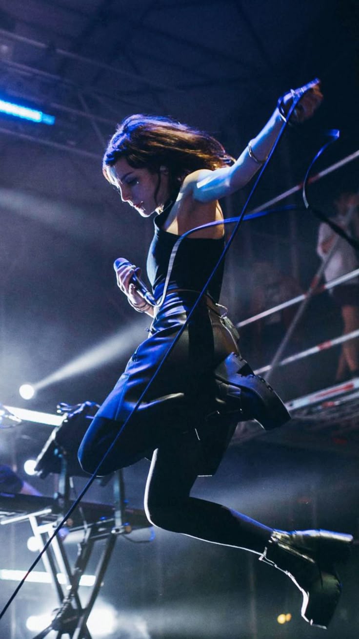 a woman is performing on a stage, singing into a microphone. she is jumping in the air and is wearing a dark dress, dark tights, and platform boots. she has dark hair. the stage is lit with spotlights. a wire runs from the microphone. an instrument is visible on the stage. there is an audience in the background.