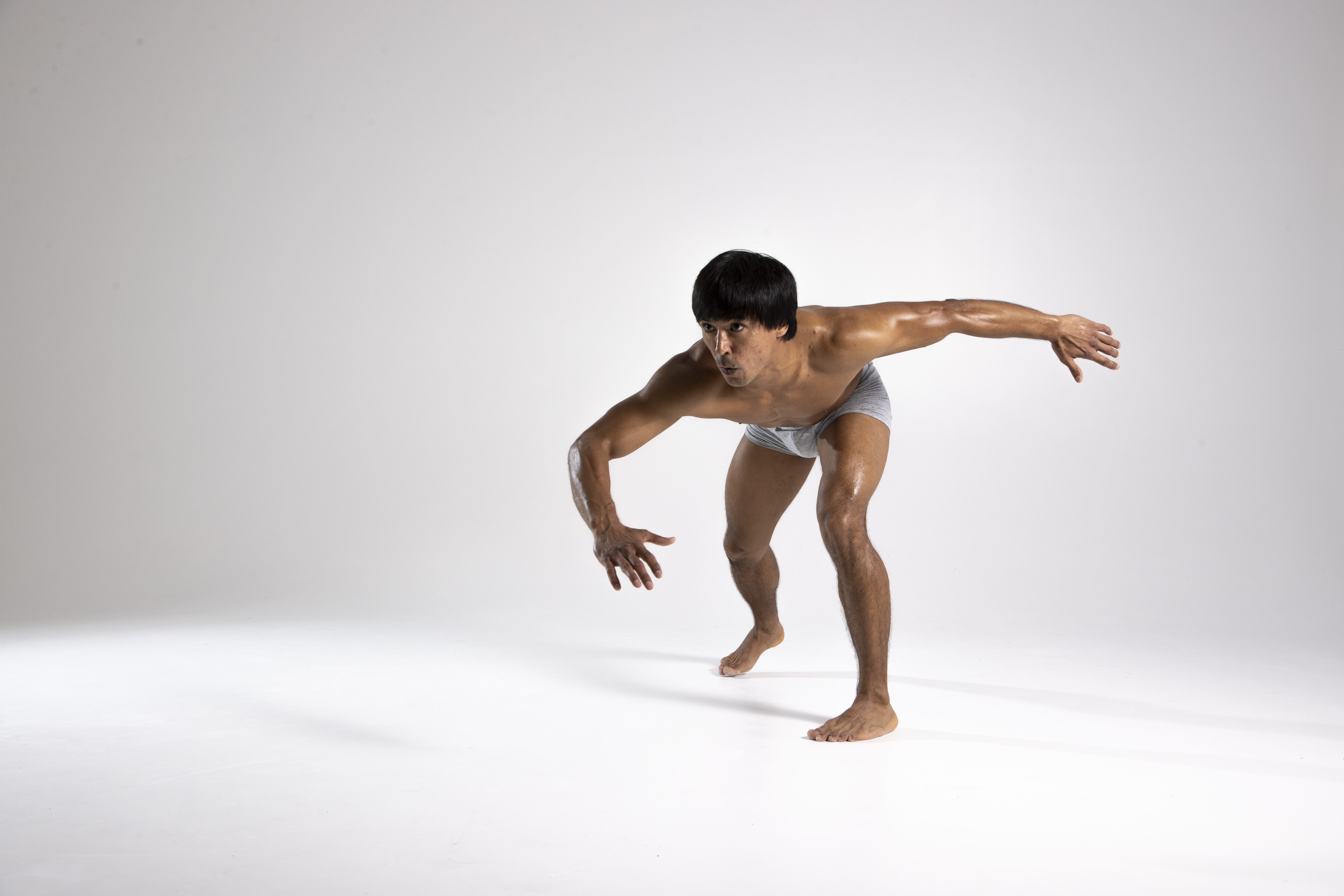 a muscular, oiledskinned man is crouching forward in a ready stance against a white background. he has short dark hair and is wearing gray underwear. his arms are extended out to the sides, hands slightly curved. he appears focused and determined, possibly depicting a martial artist.