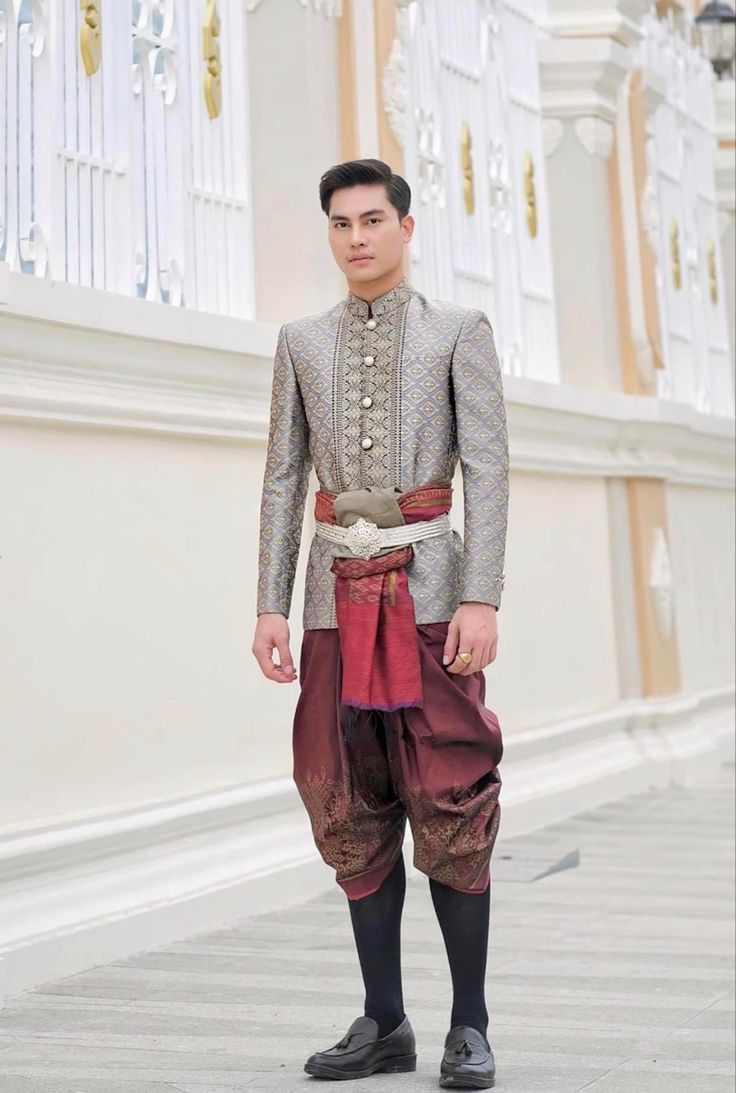 a man is dressed in a traditional thai wedding outfit. he is wearing a longsleeved jacket with gold embroidery, a gold belt with a silver buckle, a red sash, and maroon pants with gold embroidery. he is standing in front of a white building with a white, ornate railing. he is smiling and looking at the camera. the image is taken from the waist up.  he is wearing black loafers and black socks.  the image is taken in a wide shot.