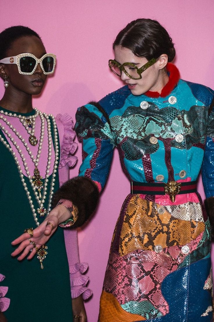 two models stand close together against a bright pink background. the model on the left wears a dark teal dress with pink frills at the shoulders and cuffs. she has on a layered necklace featuring pearls and several pendants, and large, whiteframed sunglasses. the model on the right wears a vibrant, multicolored patchwork coat made of snakeskin in various shades and patterns.  the coat features a fur trim at the cuffs and a snakeskinpatterned belt.  both models are wearing bold sunglasses and substantial jewelry. the image appears to be a backstage shot at a fashion show, likely for gucci, given the distinctive style and use of snakeskin.
