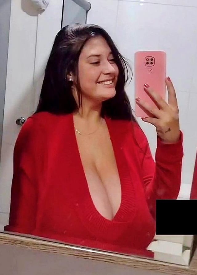a brunette woman is taking a selfie in a bathroom mirror. she is smiling and wearing a red sweater. she is leaning forward slightly and her cleavage is showing. the phone is in her right hand and she is using her left hand to hold the phone.  the bathroom is white and has white tile floors.  there is a white counter top and a mirror.