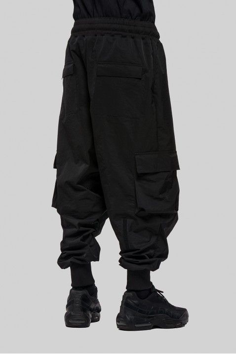 a man is wearing black cargo pants with a drawstring waist and large pockets on the legs. he is wearing black sneakers with a white sole. he is standing with his legs slightly spread apart and his arms at his side.  the pants are baggy and the sneakers are stylish. this is a casual look that is popular with men in the streetwear style.  the model is wearing a black shirt, but it is not visible in the image.