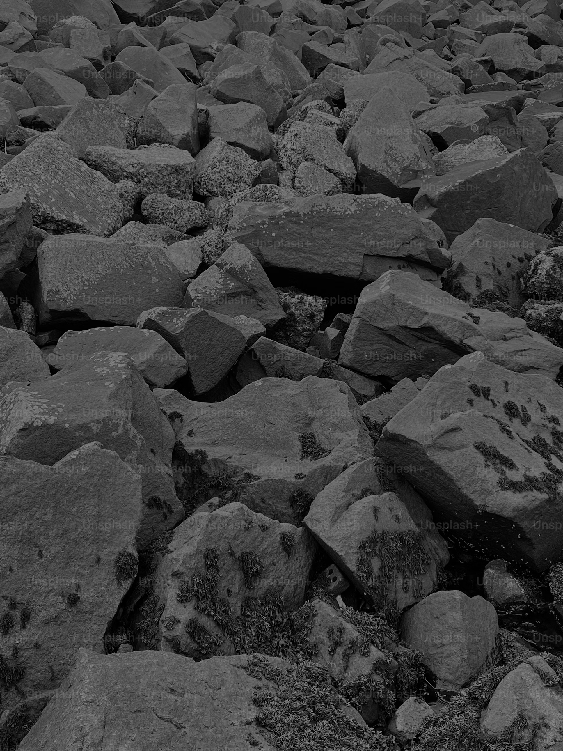 the image shows a large pile of rocks of various sizes and shapes. they appear to be dark gray or black and have a rough, uneven texture. the rocks are piled on top of each other, creating a sense of depth and weight. the image has a monochrome aesthetic, with shades of gray dominating the composition.