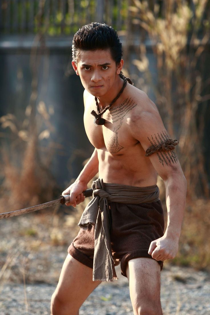 a muscular man wearing shorts and holding a sword is looking at the camera seriously. he is shirtless, has a serious expression on his face and has tattoos all over his chest and arm. he looks like a dangerous warrior ready for battle.