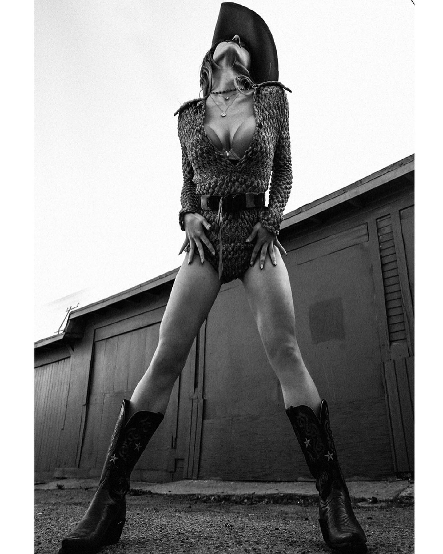 a black and white, fulllength shot of a female model striking a confident pose. she's wearing a knitted bodysuit, cowboy boots, and a cowboy hat.  her legs are spread wide, and she's looking up towards the sky. the background features a grey building with garage doors. the overall aesthetic is vintage or retroinspired, with a focus on fashion and a strong female presence.  the image is stylish and evocative.