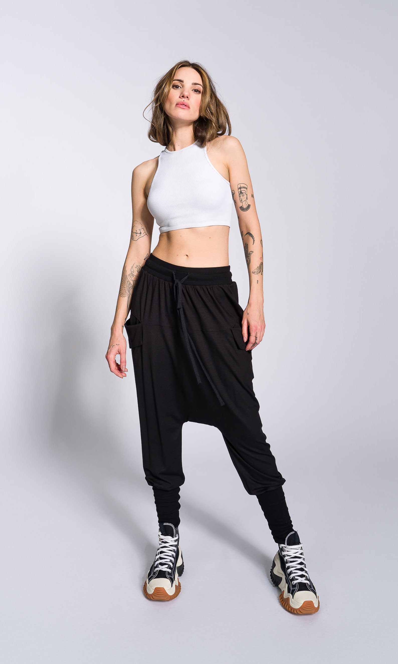 a young woman with tattoos is standing against a light grey background. she is wearing a white crop top and black harem pants with drop crotch. her footwear consists of high top sneakers. she is looking confidently towards the camera.  the overall style is casual and trendy. the image is a welllit, studio shot, showing the model from head to toe.